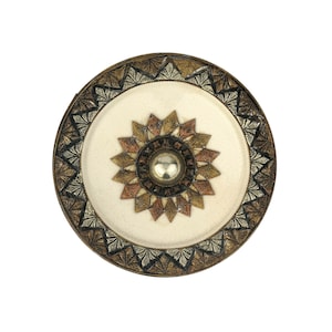 Pode incluir: Um prato redondo decorativo com um centro branco e um design geométrico preto, dourado e cobre ao redor da borda. O centro apresenta um design de estrela com um botão prateado no meio.