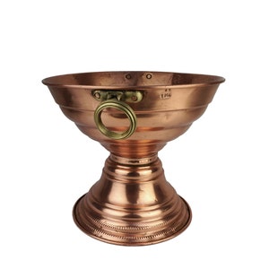 Puede incluir: Un cuenco de color cobre con base de pedestal y asa de latón. El cuenco tiene un diseño acanalado y un borde decorativo. El asa está sujeta al lateral con un anillo de latón. El cuenco es probablemente vintage, con unos 25 cm de diámetro.