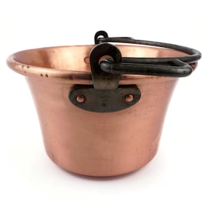 Pode incluir: Um pequeno balde cor de cobre com uma alça e suportes de metal escuro. O balde tem uma forma arredondada com uma borda ligeiramente alargada. A alça é fixada ao balde com rebites. O design geral é simples e funcional.