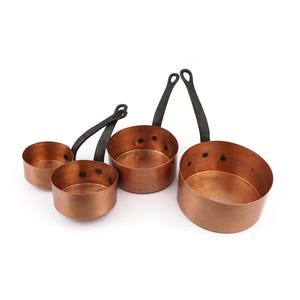 Könnte beinhalten: Vier Kupfertöpfe mit schwarzen Griffen auf weißem Hintergrund. Die Töpfe sind unterschiedlich groß und haben glänzende Kupferinnen- und Außenseiten. Die Griffe sind gebogen und mit Nieten befestigt. Die Töpfe sind zum Kochen geeignet.
