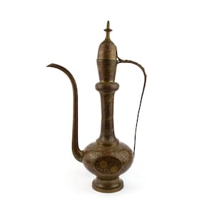 Op de afbeelding: Een antieke bronzen metalen kan met een lange, gebogen tuit en een handvat. De kan heeft ingewikkelde bloempatronen en een decoratief deksel. Het ontwerp doet denken aan Midden-Oosterse of Indiase ambacht.