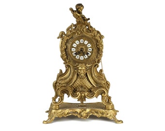 May include: A gold-toned, ornate clock with a cherub figure on top and a Roman numeral clock face. The clock has a decorative base with intricate details.