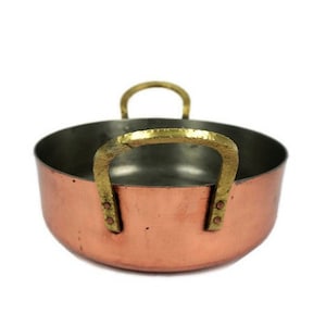 Puede incluir: Una pequeña cacerola de cobre con dos asas de latón. La cacerola tiene un acabado martillado y es ideal para cocinar en la estufa.