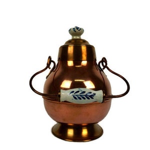 Puede incluir: Una tetera de cobre con tapa y asas de porcelana blanca y azul. La tetera tiene un cuerpo redondo y una base acampanada.