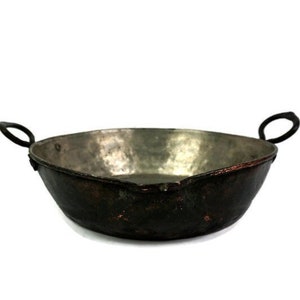 Puede incluir: Un cuenco de metal martillado con dos asas de bucle. El cuenco tiene un exterior oscuro y envejecido y un interior más claro y reflectante. Las asas son negras y curvadas. El cuenco probablemente se utiliza para cocinar o servir.