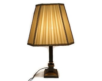 May include: A gold table lamp with a pleated beige lampshade. The lamp has a square base and a cylindrical stem.