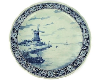 May include: A blue and white ceramic plate with a decorative border featuring a windmill scene. The plate depicts a windmill with a house and a canal in the background.