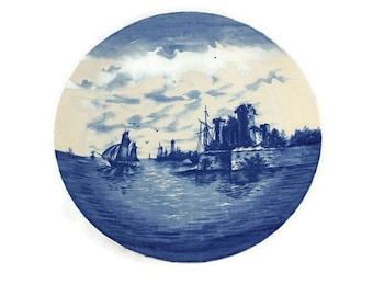 May include: A blue and white ceramic plate with a scene of ships sailing past a fortified city. The plate has a white background with a blue sky and water.