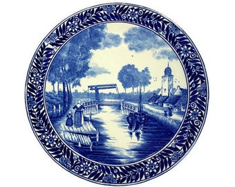 May include: A blue and white ceramic plate with a Dutch canal scene. The plate features a bridge, a church, and people in traditional clothing. The border of the plate is decorated with a floral pattern.