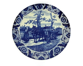 May include: A blue and white ceramic plate with a Dutch scene of a woman walking a horse pulling a cart. The plate has a floral border.