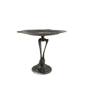 May include: A silver-colored metal serving tray with a decorative, organic design. The tray has a wide, oval-shaped top and a tall, slender base with three legs.