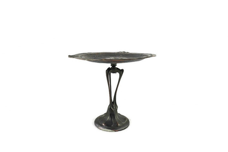 May include: A silver-colored metal serving tray with a decorative, organic design. The tray has a wide, oval-shaped top and a tall, slender base with three legs.