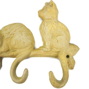 Puede incluir: Un conjunto de tres ganchos de hierro fundido amarillo con forma de gatos en una rama. Los ganchos est&aacute;n dise&ntilde;ados para colgar art&iacute;culos como abrigos, sombreros o toallas.