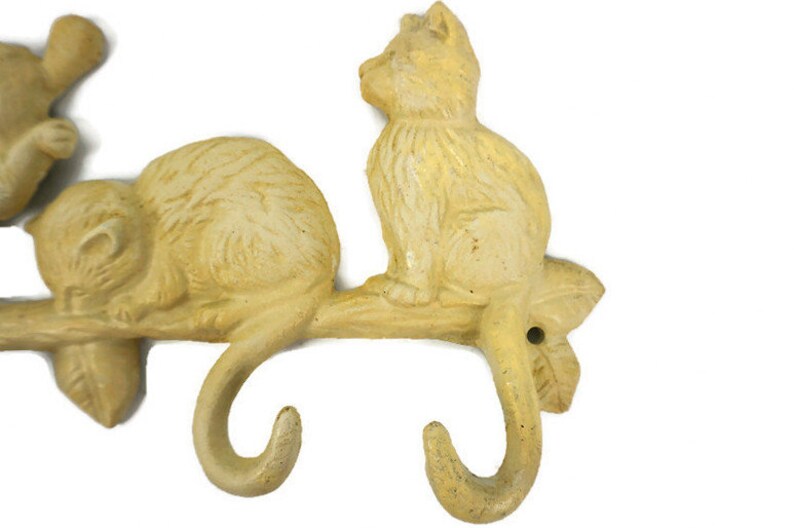 Puede incluir: Un conjunto de tres ganchos de hierro fundido amarillo con forma de gatos en una rama. Los ganchos est&aacute;n dise&ntilde;ados para colgar art&iacute;culos como abrigos, sombreros o toallas.