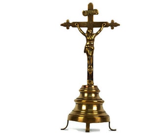 May include: A gold-toned metal crucifix with a figure of Jesus Christ on the cross. The crucifix is mounted on a decorative stand with a round base and three legs.
