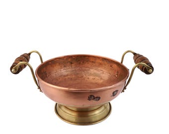 May include: A copper bowl with brass detailing and wooden handles. The bowl is round and rests on a brass base. The handles are made of wood and are attached to the bowl with brass supports.