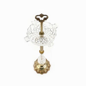 Op de afbeelding: Een vintage asbak met een heldere, diamantgeslepen glazen kom en een messing standaard. De standaard heeft een decoratieve basis en een handvat aan de bovenkant. De asbak is ontworpen voor sigaretten en sigaren.