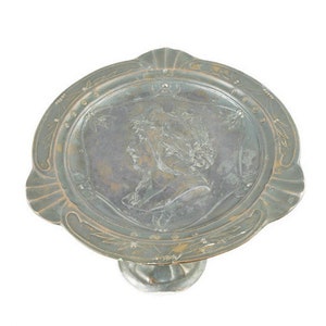 May include: A silver-toned metal cake stand with a raised, embossed design of a woman's head. The stand has a round top and a single, ornate foot.