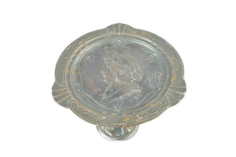 May include: A silver-toned metal cake stand with a raised, embossed design of a woman's head. The stand has a round top and a single, ornate foot.