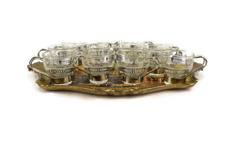 May include: A set of 10 clear glass cups with silver handles and etched designs, arranged on a gold-coloured tray with a floral pattern.