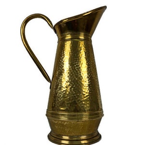 Könnte beinhalten: Eine gehämmerte Messingkanne mit einer breiten Öffnung und einem geschwungenen Griff. Die Kanne hat eine strukturierte Oberfläche und eine goldene Oberfläche.