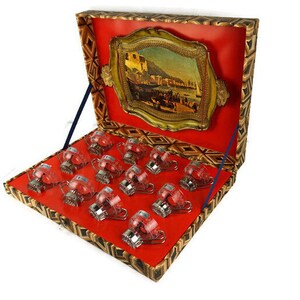 May include: A set of 12 clear glass cups with silver accents, arranged in a red and gold patterned box. The box has a hinged lid with a gold-framed picture of a cityscape.