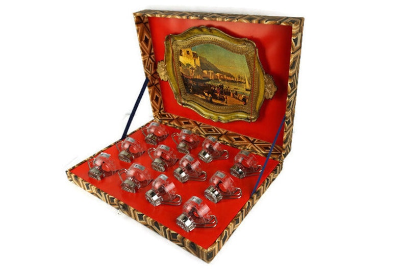 May include: A set of 12 clear glass cups with silver accents, arranged in a red and gold patterned box. The box has a hinged lid with a gold-framed picture of a cityscape.