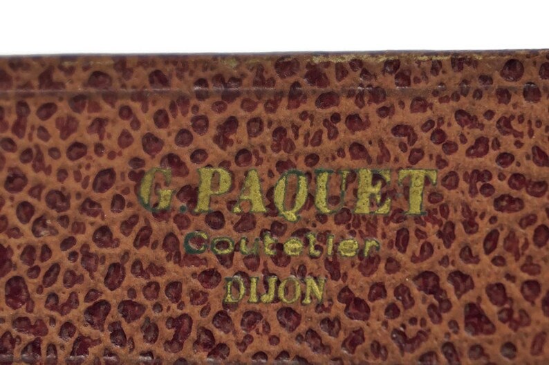 May include: Close-up of a brown leather box with gold lettering that reads "G. Paquet Couteller Dijon".