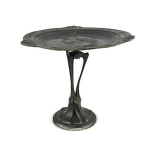 May include: A silver-colored metal serving tray with a raised design on the rim and a stylized figure in the center. The tray is attached to a pedestal with a unique, organic design.