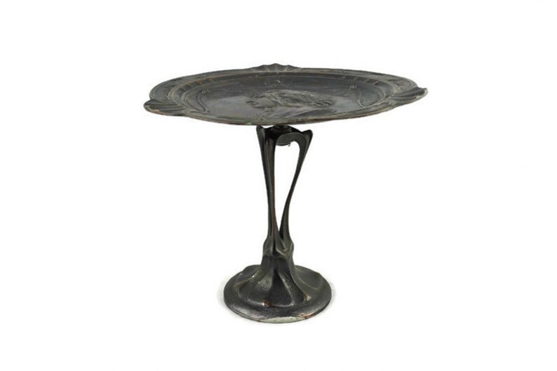 May include: A silver-colored metal serving tray with a raised design on the rim and a stylized figure in the center. The tray is attached to a pedestal with a unique, organic design.