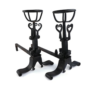 Puede incluir: Par de hacheros de chimenea de hierro forjado negro. Cada hachero presenta una base curva, una columna central con adornos decorativos y una parte superior con un asa curva. Los hacheros están diseñados para sostener leños en una chimenea.