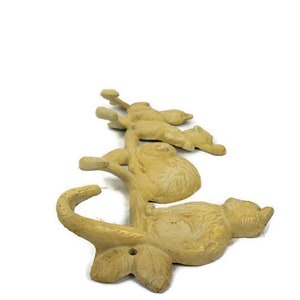 Puede incluir: Un perchero de pared de hierro fundido con cinco ganchos con forma de gatos. Los gatos est&aacute;n en varias poses y est&aacute;n pintados de color amarillo claro.