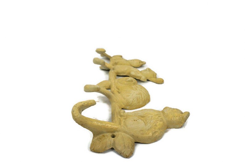 Puede incluir: Un perchero de pared de hierro fundido con cinco ganchos con forma de gatos. Los gatos est&aacute;n en varias poses y est&aacute;n pintados de color amarillo claro.