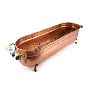 Puede incluir: Una jardinera ovalada alargada de cobre con un acabado metálico liso. Cuenta con dos asas de latón con agarres de cerámica blanca decorados con motivos florales azules. El recipiente se apoya sobre cuatro pequeñas patas de latón.
