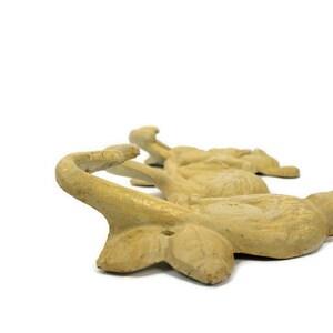 Puede incluir: Un conjunto de cuatro ganchos de hierro fundido con forma de p&aacute;jaros estilizados. Los ganchos est&aacute;n pintados de un color amarillo p&aacute;lido y est&aacute;n dispuestos en fila. Cada gancho tiene un pico curvo que forma el gancho.