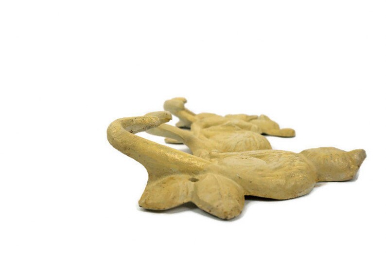 Puede incluir: Un conjunto de cuatro ganchos de hierro fundido con forma de p&aacute;jaros estilizados. Los ganchos est&aacute;n pintados de un color amarillo p&aacute;lido y est&aacute;n dispuestos en fila. Cada gancho tiene un pico curvo que forma el gancho.
