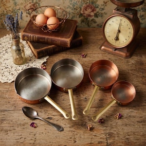 Puede incluir: Una colección de cacerolas con fondo de cobre y asas color crema, junto con una balanza antigua, libros antiguos y un pequeño jarrón de cristal con lavanda. También hay una cuchara vintage y una cesta de alambre con huevos.