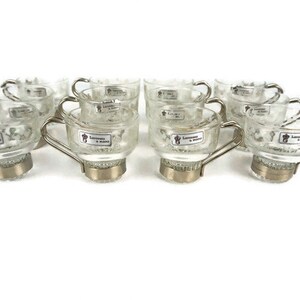 May include: Set of ten clear glass espresso cups with silver rims and handles. Each cup has a small label that reads "Lavazza Espresso & Crema".