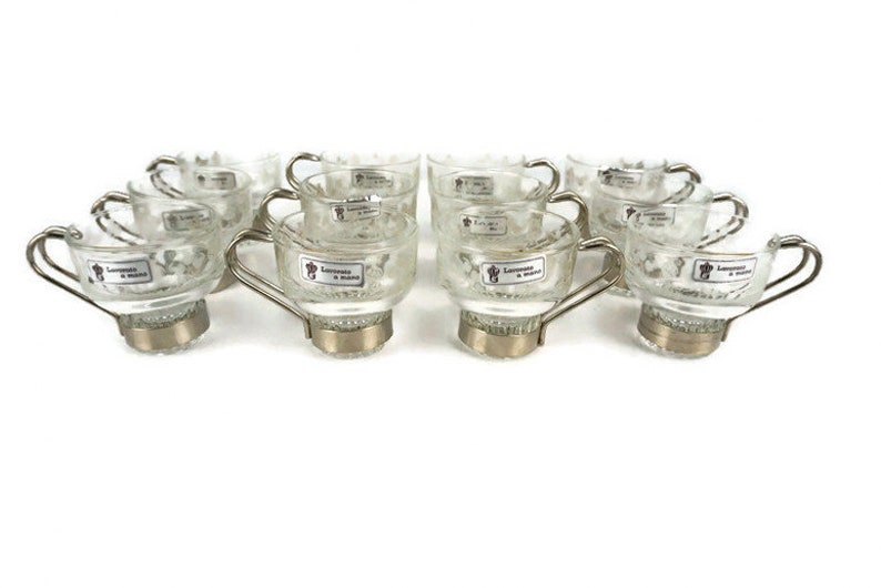 May include: Set of ten clear glass espresso cups with silver rims and handles. Each cup has a small label that reads "Lavazza Espresso & Crema".