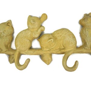 Puede incluir: Un colgador de llaves de hierro fundido amarillo con forma de rama con cuatro gatos. Dos gatos est&aacute;n sentados en la rama, uno est&aacute; durmiendo en la rama y uno est&aacute; colgado de la rama. La rama tiene tres ganchos para colgar llaves.
