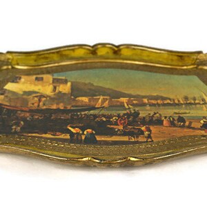 May include: A gold-coloured tray with a raised edge and a painted scene of a harbour with boats and people. The tray has a yellow-gold finish and a decorative design around the edge.