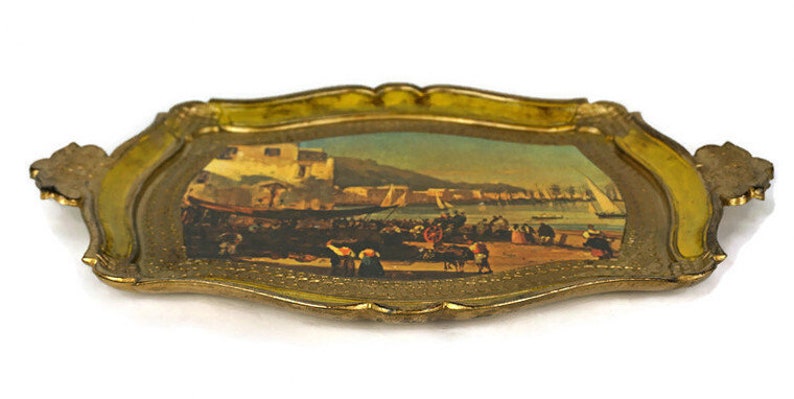 May include: A gold-coloured tray with a raised edge and a painted scene of a harbour with boats and people. The tray has a yellow-gold finish and a decorative design around the edge.