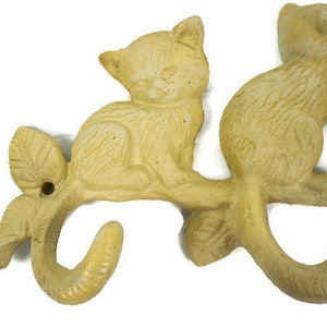 Puede incluir: Un colgador de llaves de hierro fundido con forma de rama con tres gatos sentados en &eacute;l. Los gatos y la rama est&aacute;n pintados de un color amarillo p&aacute;lido.