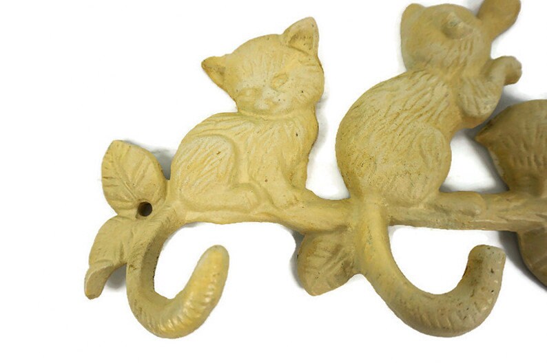 Puede incluir: Un colgador de llaves de hierro fundido con forma de rama con tres gatos sentados en &eacute;l. Los gatos y la rama est&aacute;n pintados de un color amarillo p&aacute;lido.
