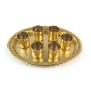 Op de afbeelding: Een goudkleurige metalen serveerschaal met zes kleine bijpassende bekers. De ovale schaal en bekers hebben decoratieve geëtste patronen. De bekers zijn in twee rijen op de schaal gerangschikt.