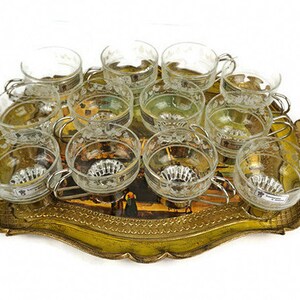 May include: A set of 12 clear glass cups with etched designs, arranged on a gold-coloured metal tray with a painted scene. The tray has a scalloped edge and ornate details.