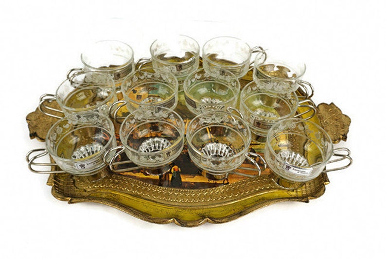 May include: A set of 12 clear glass cups with etched designs, arranged on a gold-coloured metal tray with a painted scene. The tray has a scalloped edge and ornate details.