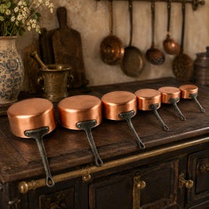 May include: A set of six graduated copper saucepans with dark, textured metal handles arranged in a row. Each pan features a polished, round copper body and a sturdy, riveted handle, displayed in descending order of size from left to right.