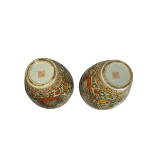 May include: Two white ceramic vases with a floral pattern in shades of orange, yellow, and green. The vases have a gold trim and a red seal on the bottom.
