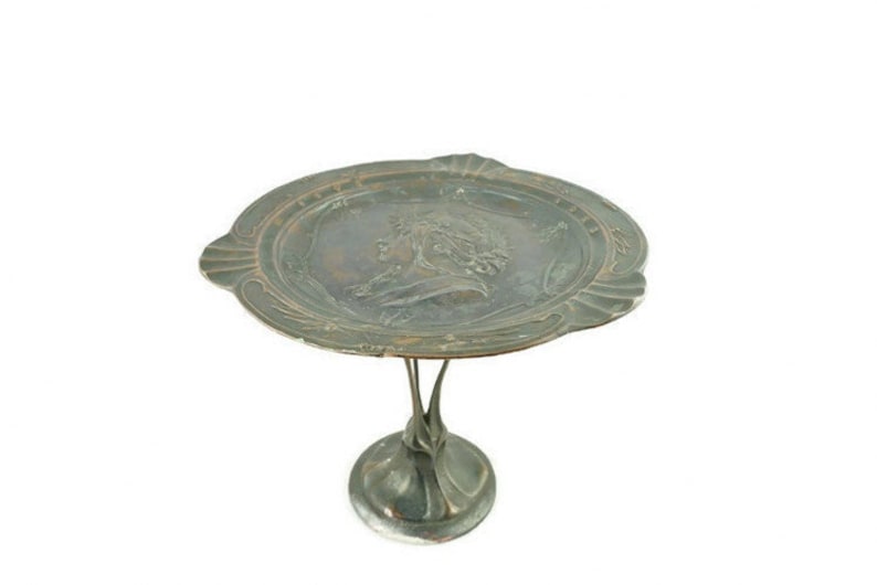 May include: A silver-toned metal cake stand with a round, decorative top featuring a raised, embossed design of a woman's profile. The stand has a delicate, ornate base.
